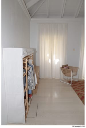 This is a photograph of a bright, minimalist interior space featuring a bedroom or dressing area with white walls, exposed ceiling beams, natural light from floor-to-ceiling curtains, and modest furnishings including a wooden clothing rack and wicker
