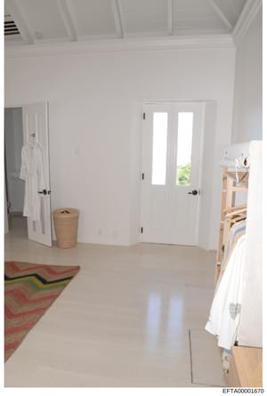 This is a photograph of a bright, minimalist bedroom interior featuring white walls, coffered ceiling, white double doors with glass panels, and wooden furniture. The room appears to be part of a residential property and may have been documented as e