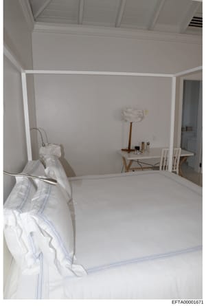 This is a photograph of a bedroom interior, featuring a four-poster canopy bed with white linens and blue trim, a wooden desk with chair, and minimalist furnishings. The room has white walls, coffered ceiling details, and appears to be part of a resi