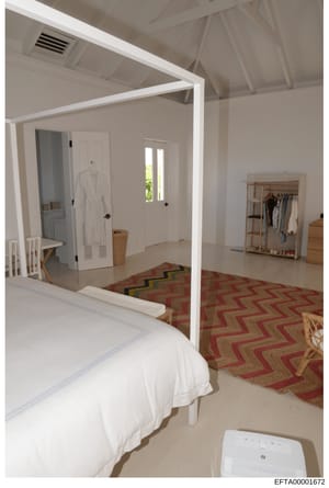 This photograph shows a modern master bedroom with an attached bathroom viewed through an open white-framed doorway. The bedroom features a four-poster bed with white linens, wooden furnishings, a red and gold geometric-patterned area rug, and a wood