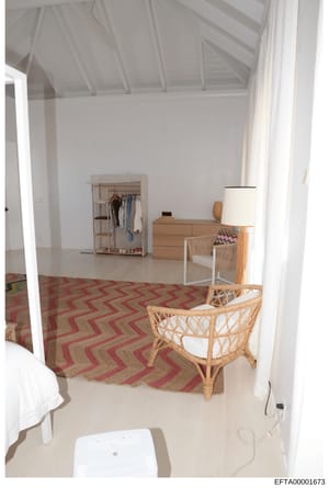 This is a photograph of an interior bedroom space showing contemporary furnishings including wooden clothing racks, dressers, rattan chairs, and a geometric patterned rug. The room features high white ceilings with exposed beams and minimalist design