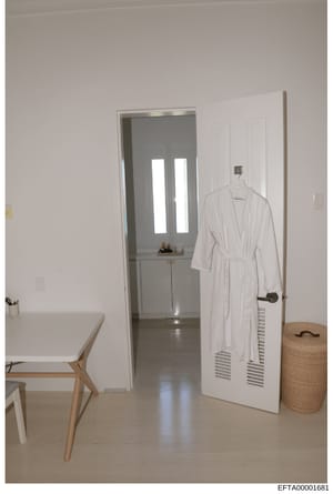 This is a photograph of an interior residential space showing a modern, minimalist bedroom or suite. The image depicts a white-walled room with a small desk area on the left, an open doorway leading to a bathroom with modern fixtures, a white robe ha