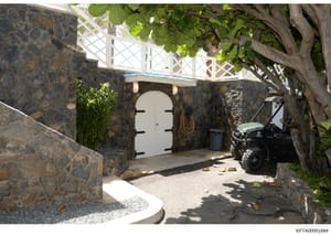 This photograph documents the exterior entrance and gated courtyard area of a property, featuring a white metal gate set into dark stone walls, with vehicle access visible. The image appears to be part of property documentation or evidence photograph
