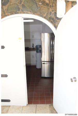 This is a photograph of a residential kitchen interior, taken through an open doorway. The image shows stainless steel refrigerator and appliances, white cabinetry, dark tile flooring, and decorative stone or marble wall accents. The photo appears to