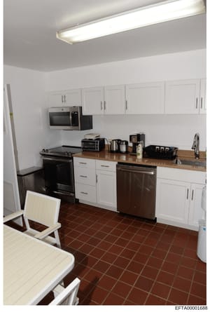 This is a photograph of a modern kitchen interior featuring white cabinetry, stainless steel appliances, and brown tile flooring. The image appears to be evidence documentation from the Epstein investigation, likely taken as part of property inventor