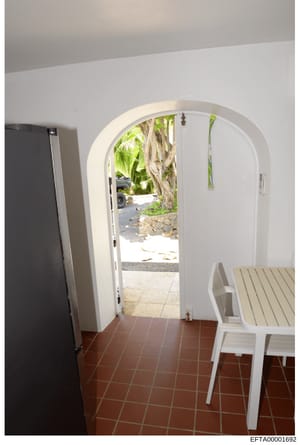 This is a photograph of an interior residential space showing a white arched doorway leading to an outdoor courtyard area. The image captures modern minimalist interior design with terracotta tile flooring, white walls, a dark refrigerator on the lef
