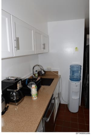 This is a photograph of a kitchen interior showing white wall cabinets, a beige countertop with various appliances including a coffee maker and water cooler, and basic kitchen fixtures. The image appears to be documentary evidence photography, likely