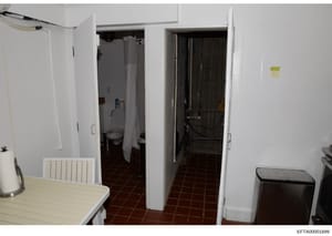 This is an evidence photograph showing the interior of a kitchen or utility area with multiple doorways leading to a bathroom and storage/mechanical room. The image depicts white walls, red-brown tile flooring, basic appliances and furnishings, and a