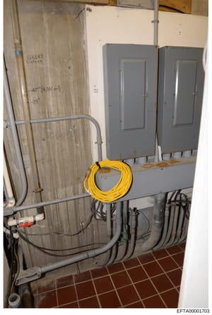 This is an evidence photograph showing electrical infrastructure in what appears to be a basement or utility area of an Epstein property. The image documents two gray electrical panels, extensive wiring, a coiled yellow extension cord, and handwritte