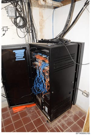 This photograph shows a black server rack or network equipment cabinet with open panels revealing extensive internal cabling and networking hardware. Blue and orange cables are organized and run through the interior framework. The cabinet is located 