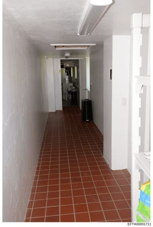 This is a photograph showing a long, narrow hallway with red/brown tile flooring and white walls. The hallway contains several doors on both sides, fluorescent lighting, and appears to be part of a commercial or residential building. The image is lik