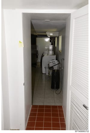 This is an evidence photograph showing a narrow utility or laundry room space, likely from one of the properties under investigation. The room contains basic appliances and storage, with red tile flooring at the entrance transitioning to gray tile. T