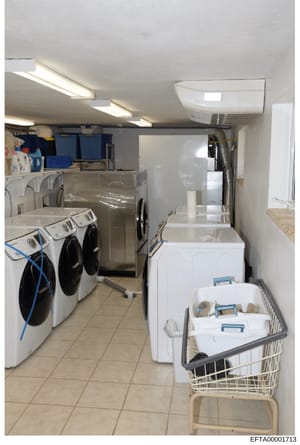 This is a photograph of a commercial or institutional laundry room showing multiple washing machines, dryers, and related equipment. The space contains white front-loading washers with blue hoses, white dryers, storage shelves with blue containers, a