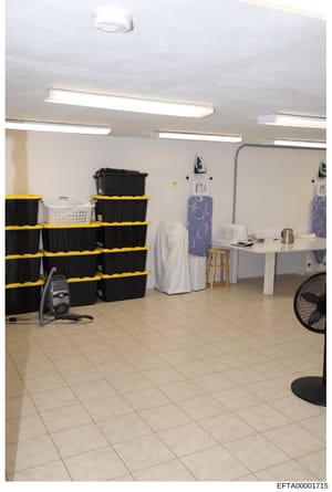 This is an evidence photograph showing the interior of what appears to be a storage, laundry, or utility room. The space contains multiple black plastic storage containers stacked with yellow banding, fluorescent lighting, mirrors, ironing boards, an