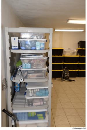 This photograph shows a tall white shelving unit filled with various household items, cleaning supplies, and storage containers. The image appears to be taken in a utility or storage room, with tile flooring and fluorescent lighting visible. The shel