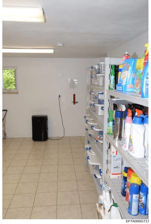 This is an evidence photograph showing the interior of a storage or utility room, likely at one of Epstein's properties. The room contains shelving units stocked with cleaning supplies, household products, and various containers. The photo documents 