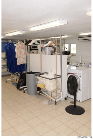 This photograph shows an institutional or residential laundry facility with commercial-grade equipment, clothing storage systems, and utility appliances. The room appears to be part of a larger household or facility property, likely documented as evi