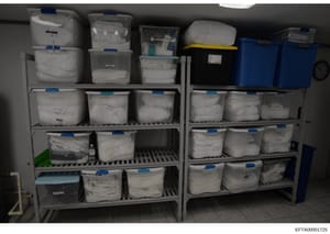 This photograph shows an organized storage facility with multiple shelving units containing numerous plastic storage containers and bins, many labeled with blue tags and labels. The containers appear to be evidence storage or archival storage, consis