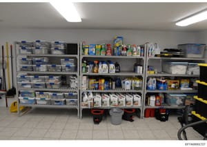 This photograph shows a well-organized storage area containing multiple shelves stocked with cleaning supplies, household products, and maintenance materials. The image captures what appears to be a utility or housekeeping storage room, likely from o