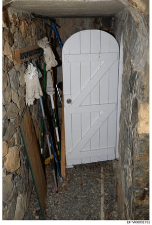 This photograph shows a narrow interior space, likely a storage closet or utility room, containing a white paneled door and various cleaning or maintenance equipment including mops, brooms, and colored cords/cables. The space has stone or concrete wa