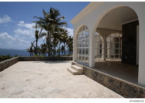 Photograph of a luxurious oceanfront property featuring a white-walled terrace with arched architectural elements, multiple floor-to-ceiling windows, and panoramic ocean views. Palm trees frame the scene, and the property appears to be located in a t