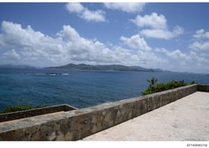 This is a photograph showing a waterfront property with a stone-walled terrace or balcony overlooking a tropical or subtropical ocean vista. The image depicts calm blue waters, distant islands with vegetation, and clear sky with white clouds. The sto