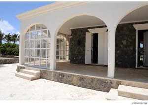 This photograph shows the exterior of a contemporary residential structure featuring clean white walls, multiple arched openings with grid-pattern windows and doors, and stone foundation elements. The property appears to be in a tropical or warm clim
