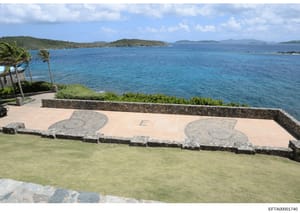 This is a photograph of a scenic oceanfront property featuring a large stone patio with decorative inlaid designs, manicured lawn, and expansive views of turquoise waters and distant islands. The image appears to be documentation of a residential or 