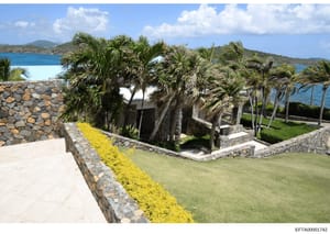 This photograph depicts a luxury coastal property featuring tropical landscaping, stone construction, and panoramic ocean views. The image shows manicured grounds with palm trees, stone walls with yellow flowering plants, and what appears to be a res