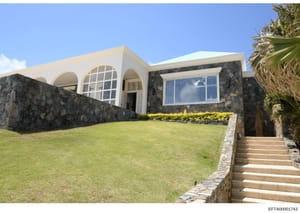 This is a photograph of a contemporary single-story residential property featuring modern architectural design with curved white walls, stone foundation, manicured landscaping, and palm trees. The image shows the front facade and grounds of what appe