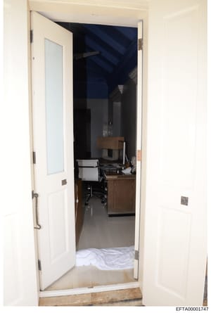 This is a photograph of a room interior taken through an open doorway, showing a workspace with a wooden desk, office chair, and various equipment. The room features cream-colored walls, a dark blue accent wall or structure visible in the background,