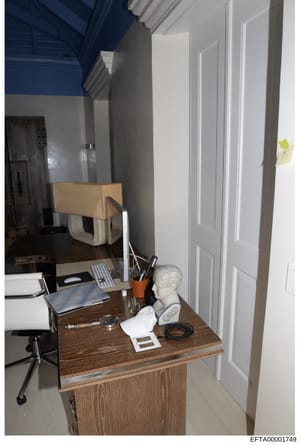 This is an evidence photograph from an Epstein property showing an interior room with a desk, office equipment, and personal items. The image captures a work or study space with contemporary furnishings, including a wooden desk, computer keyboard, an