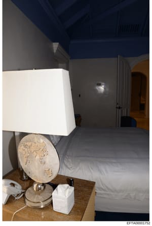 Photograph of an interior bedroom space showing a bed with gray bedding, a wooden nightstand with decorative lamp and tissue box, and walls with two-tone paint (gray lower, dark blue upper). The image appears to be documentation of a property's inter