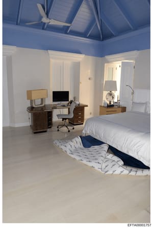This is a photograph of a modern bedroom featuring a vaulted ceiling with blue paint and white trim, white walls, wood furnishings, and a dedicated work area with computer desk. The room contains a queen-sized bed with white bedding and patterned thr