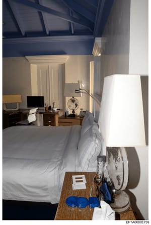 This is a photograph of a luxury bedroom interior, showing a bed with white linens, work desk with computer monitor, and various furnishings. The room features high ceilings with blue beam work, cream-colored walls, and upscale decorative elements. T