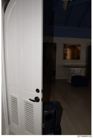This is an evidence photograph showing the interior of a room with a white double-louvered door open to reveal an adjoining space. A dark piece of equipment (possibly audio/surveillance equipment) is visible in the foreground. The photograph appears 