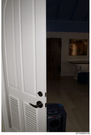 This is an evidence photograph showing an interior doorway with white louvered doors, black hardware, and a view into an adjacent room. The photo captures interior architectural details and furnishings, likely documented as part of property evidence 
