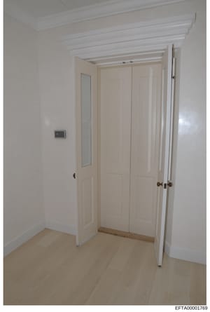 This is a color photograph of an interior hallway or corridor, showing white walls, exposed beam ceiling details, cream-colored closet or wardrobe doors with multiple panels, light wooden flooring, and a small wall-mounted switch or control panel. Th