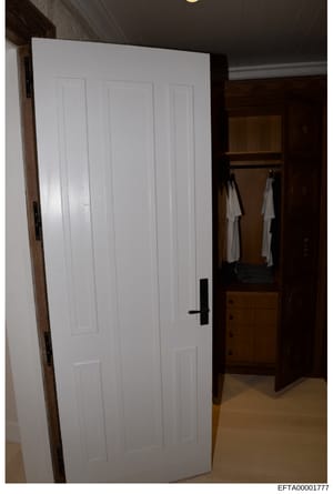 This is an evidence photograph showing the interior of a bedroom closet space with white multi-panel doors, wooden framing, and white garments hanging on a rod inside. The image appears to be part of property documentation from an Epstein investigati