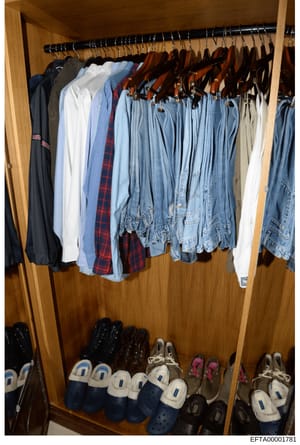 This is a photograph of a wooden closet containing various clothing items on hangers and shoes on the floor. The closet holds a collection of casual clothing including jeans, shirts, sweaters, and jackets in multiple colors, along with casual footwea