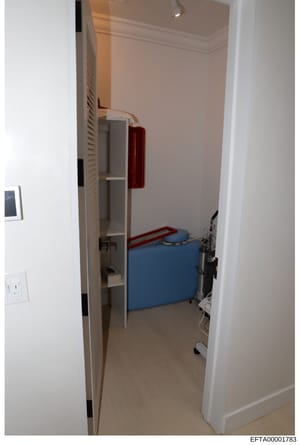 This is a photograph of a small, sparsely furnished room taken through an open doorway. The room contains a blue storage container or small bed, red metal shelving, and built-in shelving units. The photograph appears to be evidence documentation from