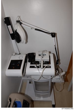 This photograph documents a collection of medical or aesthetic treatment equipment mounted on a white wall in what appears to be a residential room. The setup includes a control unit with a digital display panel, articulating arm apparatus with black