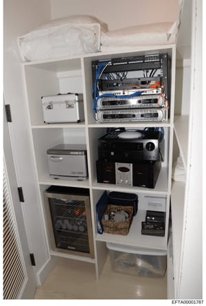 This photograph shows the interior of a white built-in cabinet containing various electronic equipment, kitchen appliances, and storage items. The cabinet appears to be part of a residential space and includes networking/audio equipment, a dishwasher