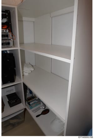 Photograph of an open white shelving unit or built-in cabinet containing electronic equipment and various items. The left side shows networking or computer hardware with orange and blue cables, while the right side shows empty white shelves. The bott