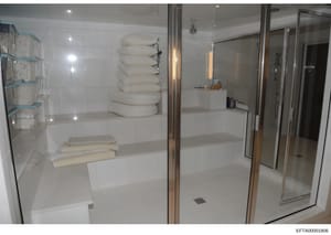This photograph depicts a high-end bathroom interior featuring a frameless glass shower enclosure with chrome fixtures, white built-in shelving with rolled towels, and a shelving unit on the left side containing folded linens. The image appears to be