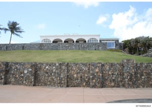 This photograph shows a contemporary residential property with distinctive architectural features, including a stone perimeter wall, manicured lawn, and a white modernist building with curved openings. The property appears to be a high-end coastal or