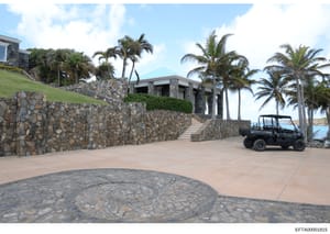This photograph shows the exterior of a large residential property featuring modern stone construction, multiple levels built into a hillside, extensive stonework walls, and tropical palm trees. The property appears to be located on a coastal area wi