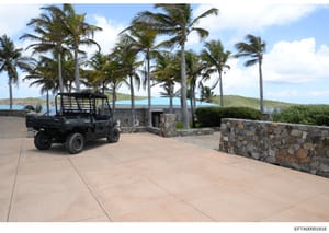 This photograph shows a tropical island property with a large paved courtyard area, native palm trees, and clear views of blue ocean water in the background. A black utility vehicle is parked in the foreground. The image documents the exterior ground