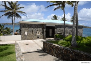 This photograph shows a modern residential structure with stone construction located on a Caribbean waterfront property. The building features a distinctive turquoise roof, stone walls, palm tree landscaping, and direct water views. The property appe