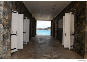 This photograph shows an interior stone corridor or covered passage with white wooden doors on both sides, leading to a bright oceanside view. The passage features dark stone walls, a wooden ceiling with recessed lighting, and opens onto a turquoise 